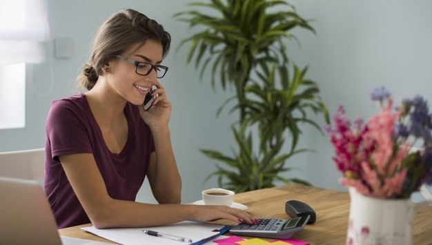 foto mulher branca usando oculos trabalhando em casa, ela toma café. na mesa de trabalho tem computador, calculadora, caneca e xícara de café