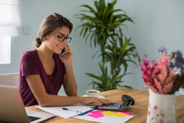 foto mulher branca usando oculos trabalhando em casa, ela toma café. na mesa de trabalho tem computador, calculadora, caneca e xícara de café