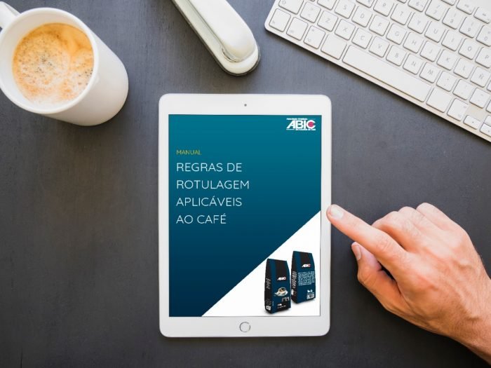 foto de uma mesa com um tablet, teclado e uma xícara de cafe. na tela do tablet a capa do manual de rotulagem aplicaveis ao café