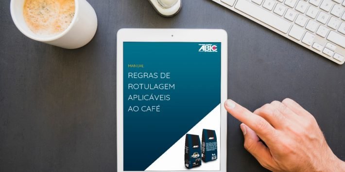 foto de uma mesa com um tablet, teclado e uma xícara de cafe. na tela do tablet a capa do manual de rotulagem aplicaveis ao café