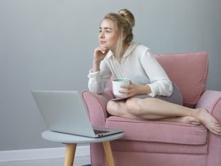 imagem mulher loira sentado no sofá rosa com xícara de café na mão olhando para computador