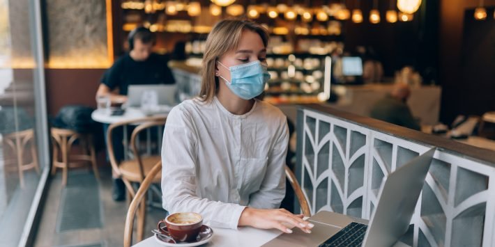 mulher loira de mascara no rosto sentada numa cafetereia usando notebook e tomando xícara de café
