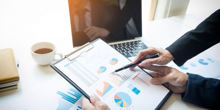 imagem de homens analisando dados em papel sobre a mesa com notebook e uma xicara de cafe