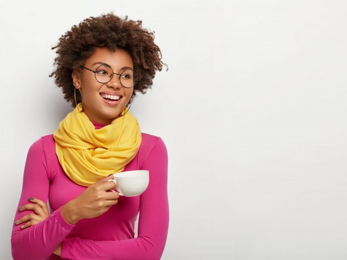 Smiling glad dark skinned woman holds mug with aromatic coffee, has pleasant talk during drinking tea, wears optical glasses, yellow scarf and pink turtleneck, isolated over white background.