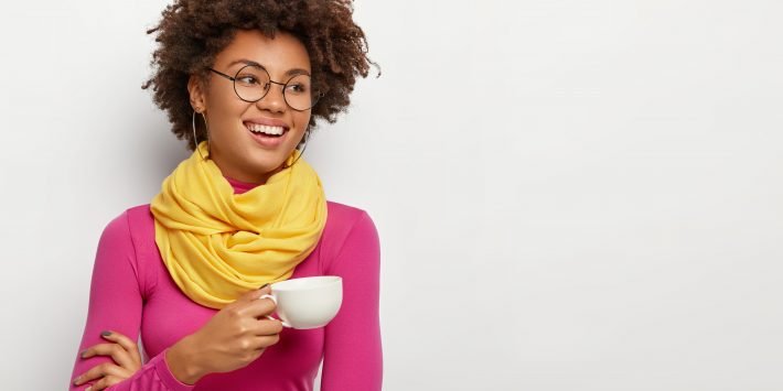 Smiling glad dark skinned woman holds mug with aromatic coffee, has pleasant talk during drinking tea, wears optical glasses, yellow scarf and pink turtleneck, isolated over white background.