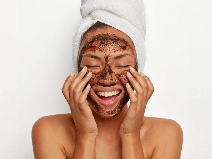 Portrait of cheerful smiling dark skinned woman applies natural coffee mask, makes circular motions with hands and massages skin, stimulates facial blood supply, wears wrapped towel on head.